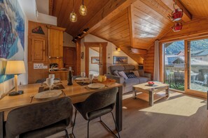 Interior acogedor de una cabaña de madera con zona de comedor, sofá y un ventanal con vistas a un paisaje montañoso.