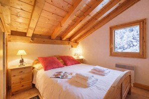 A cozy bedroom with a wooden ceiling, a bed with white bedding, red pillows, a bedside table with a lamp, and a window with a snowy view.