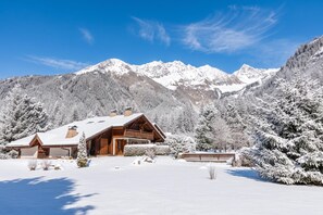 A snow-covered mountain lodge with a wooden exterior and a large balcony, surrounded by snow-laden trees and mountains.