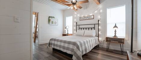 A bedroom with a bed, a ceiling fan, a desk with a lamp, and a window with a view.