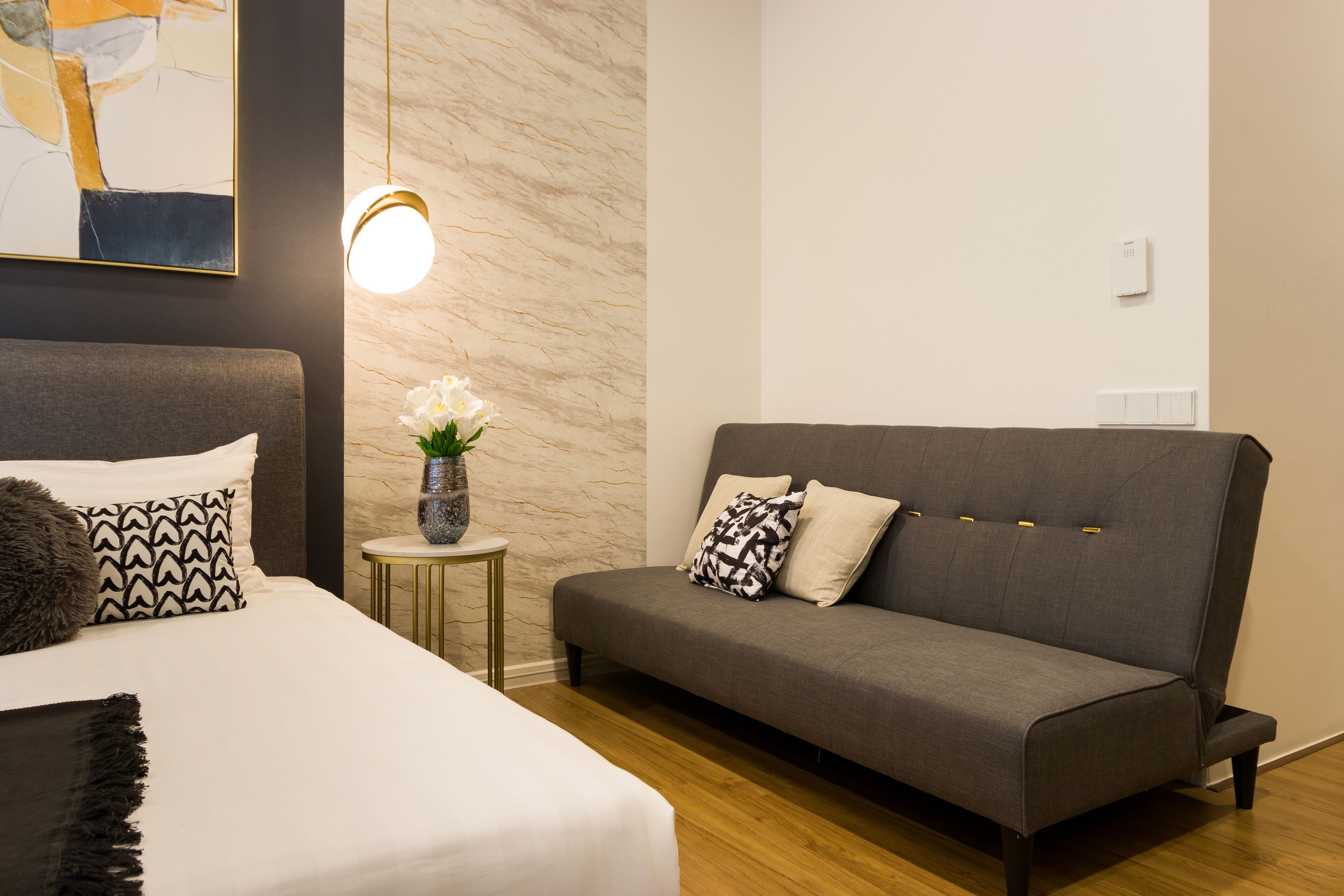 Una habitación de hotel moderna con una cama, un sofá gris, una mesita de noche con un jarrón de flores y un cuadro en la pared.