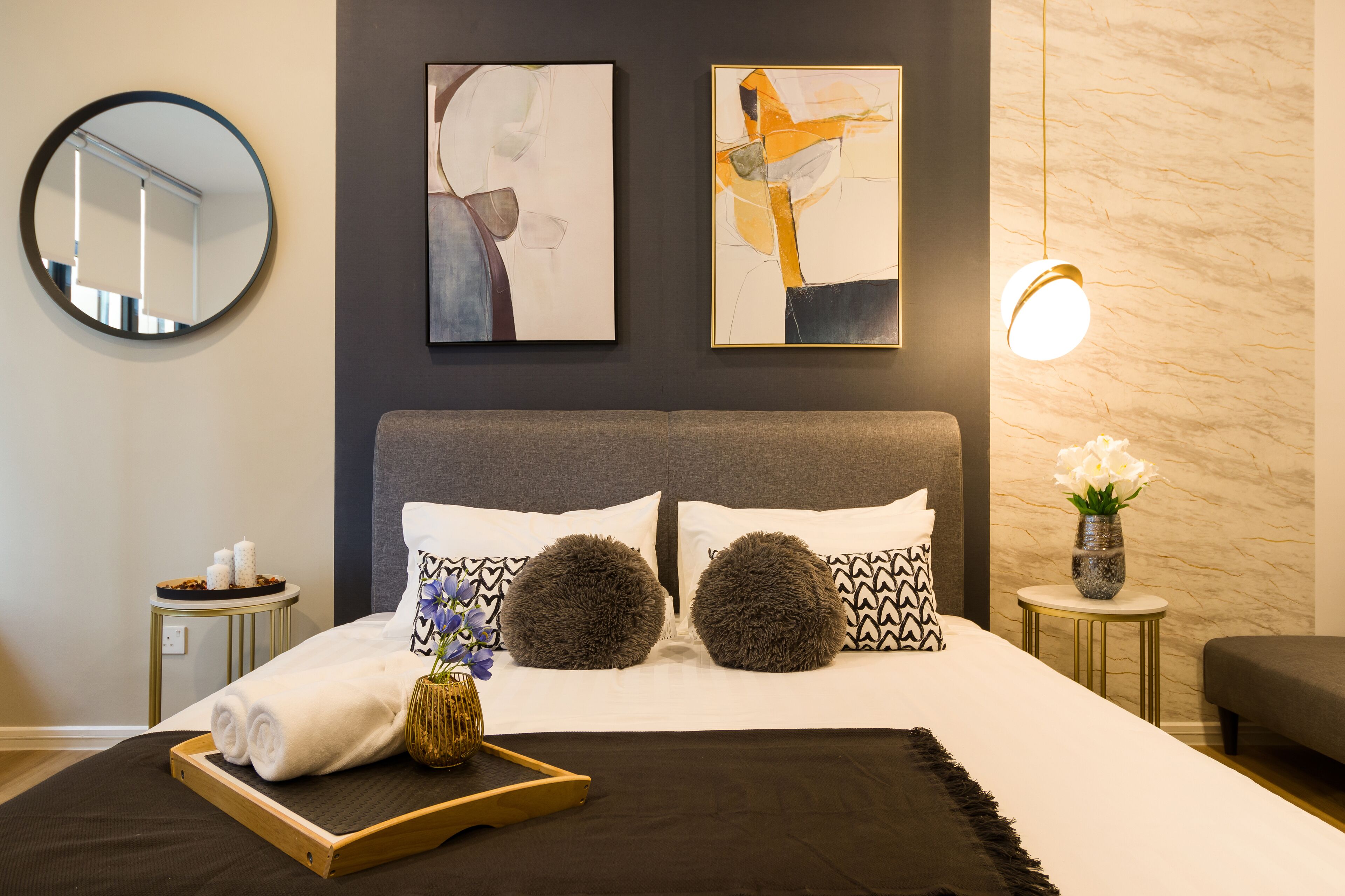 A modern hotel room with a large bed, two abstract paintings, a round mirror, a bedside table with toiletries, and a vase with flowers.