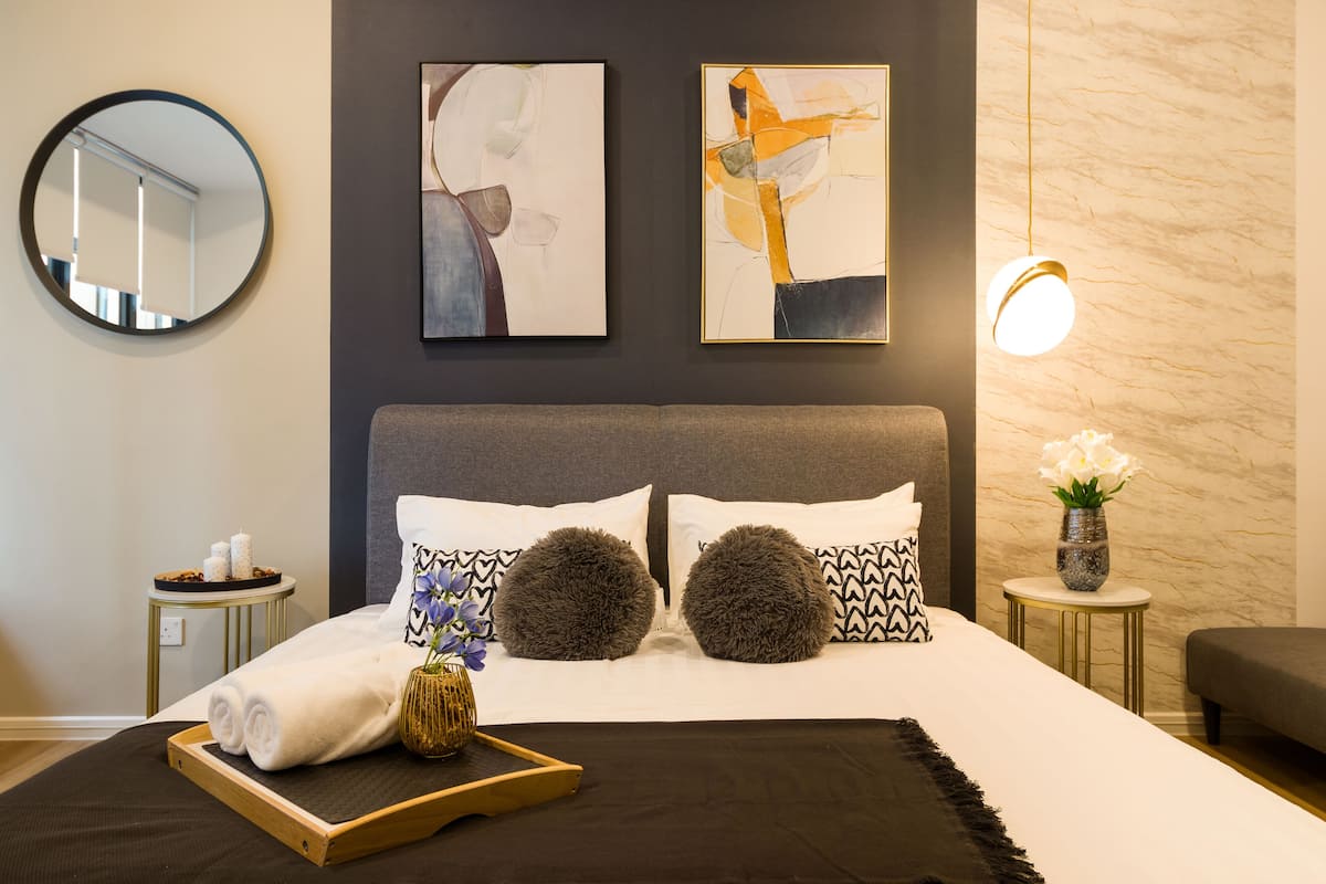 Une chambre d’hôtel moderne avec un grand lit, deux tableaux abstraits, un miroir rond, une table de chevet avec des articles de toilette et un vase rempli de fleurs.