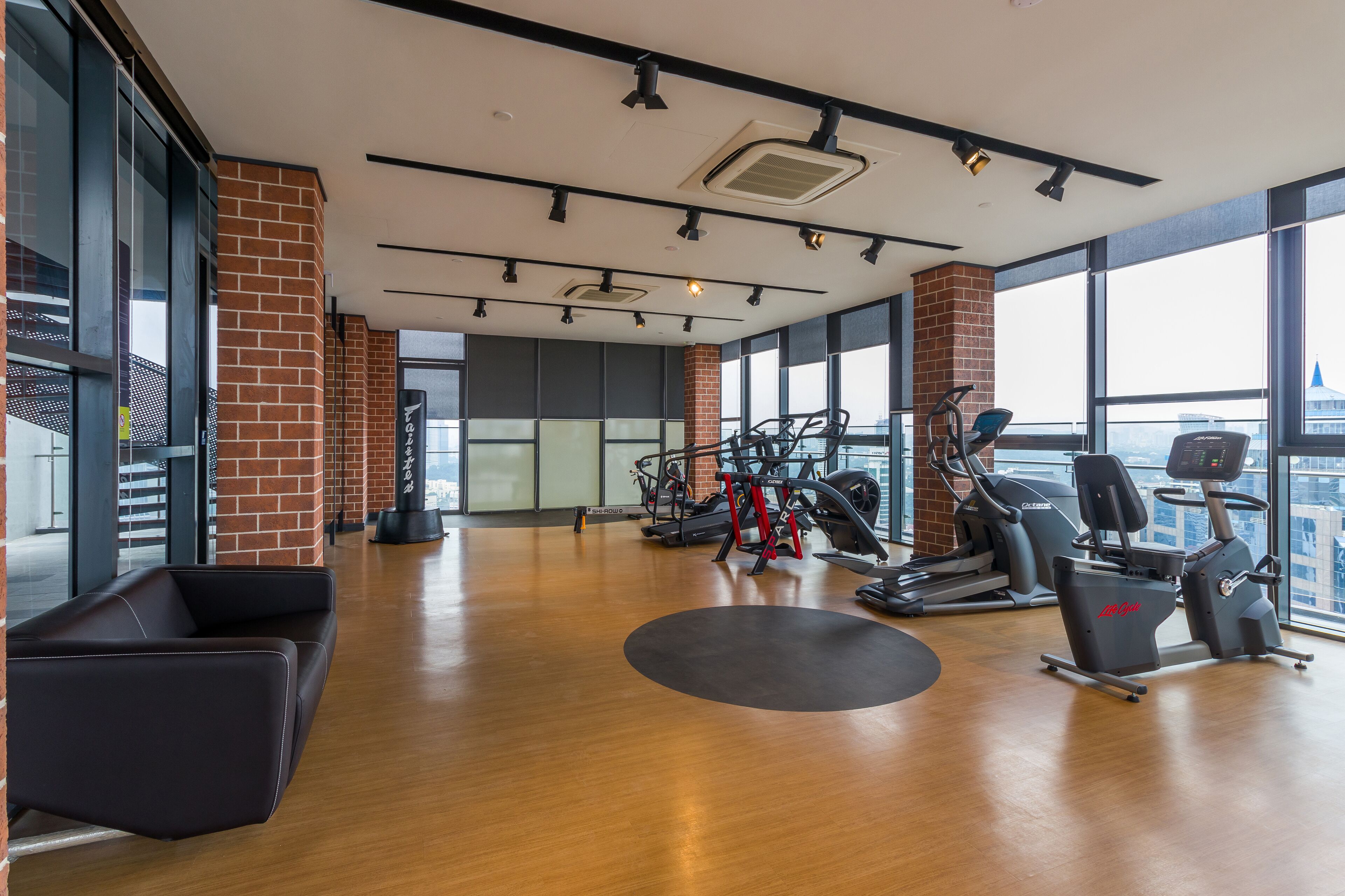 A gym with cardio machines, a black massage chair, and large windows offering a city view.