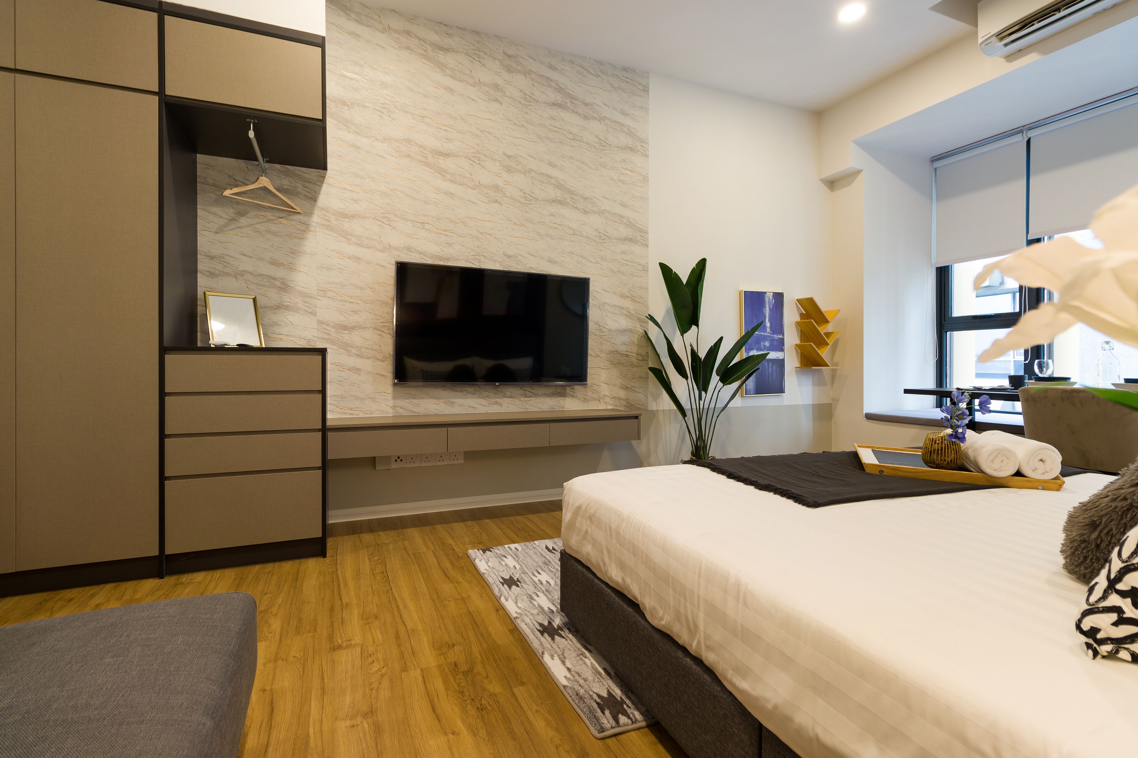 A modern hotel room with a large bed, a TV mounted on a marble wall, a wardrobe, and a small seating area.