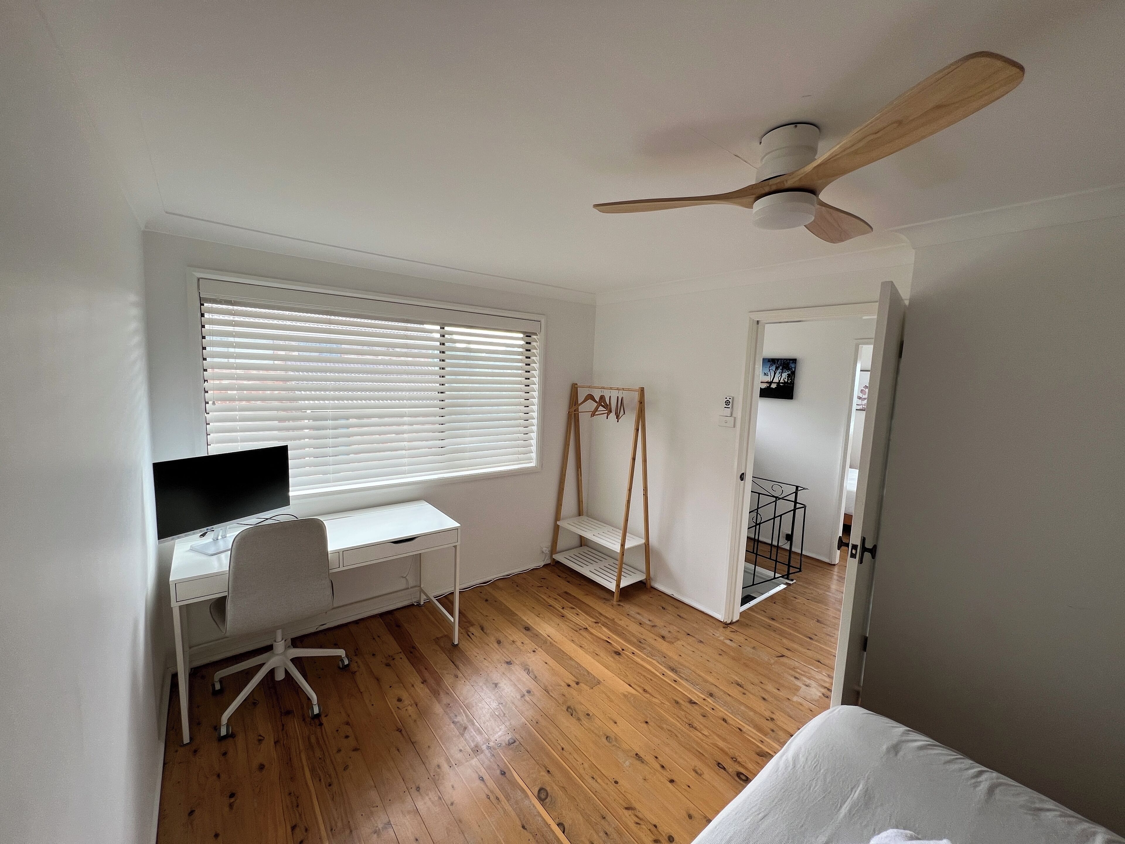 A bedroom with a wooden floor, a bed, a desk with a chair, a ceiling fan, and a window with blinds.
