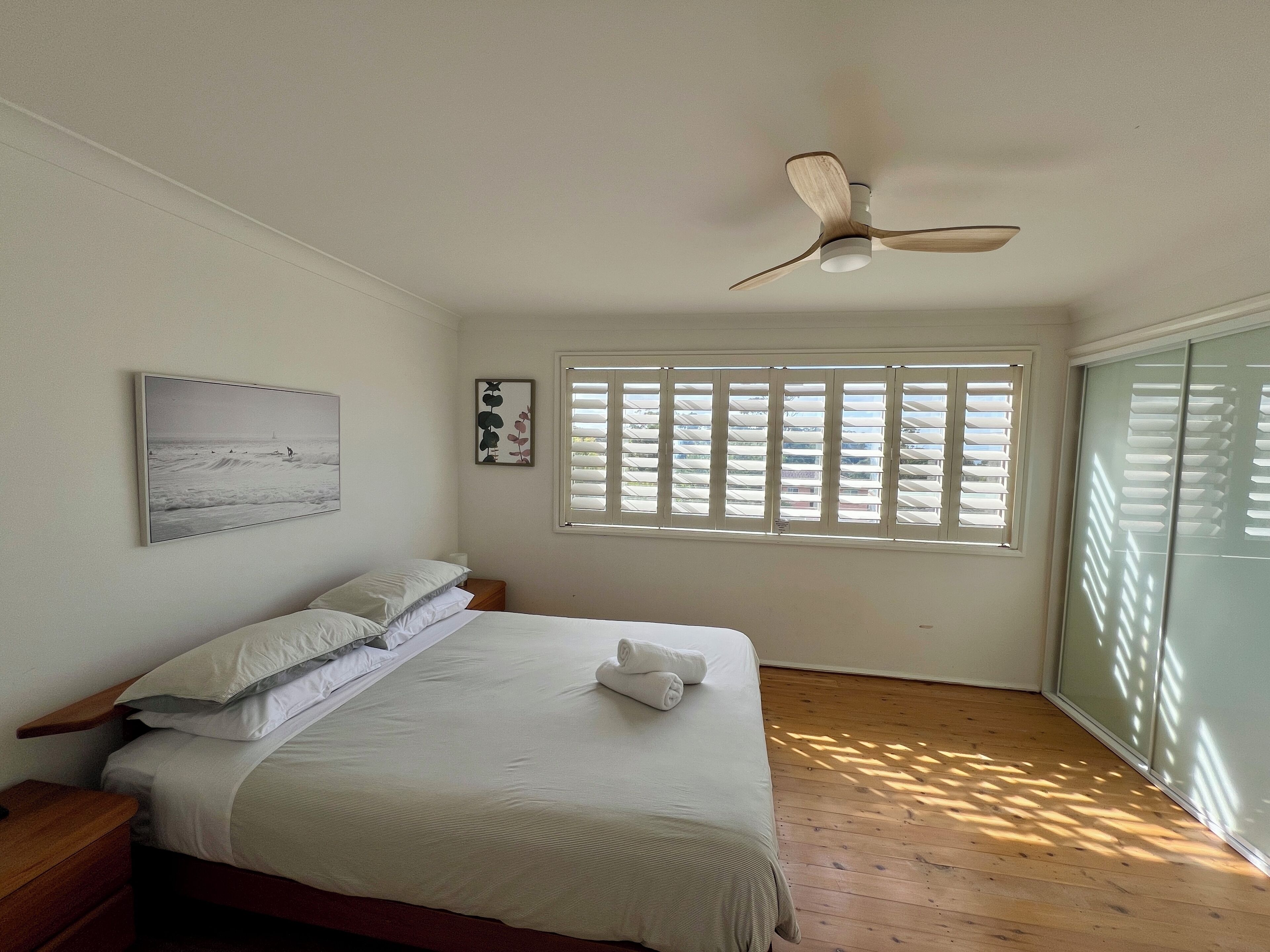 A bedroom with a bed, a ceiling fan, a window with blinds, and artwork on the wall.