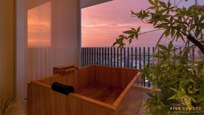 Een houten bad in een kamer met een groot raam met uitzicht op een stadsbeeld bij zonsondergang.