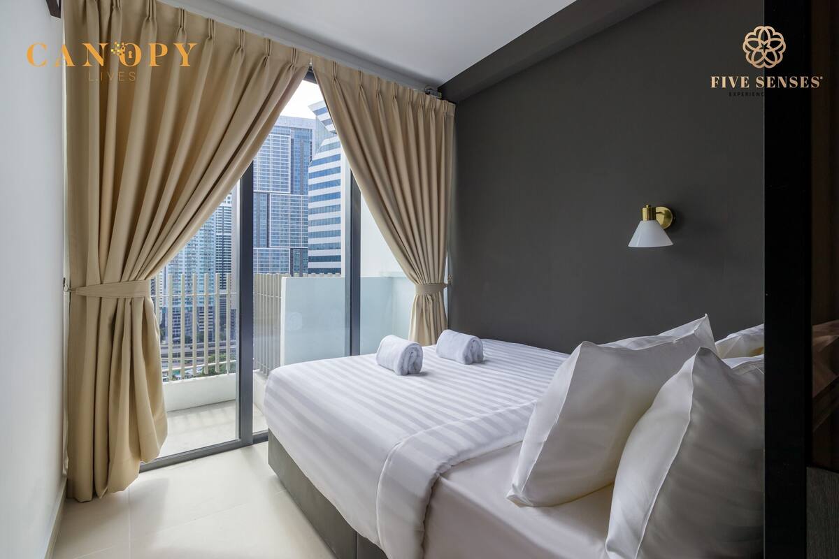 Een hotelkamer met een groot bed, twee opgerolde handdoeken, een wandlamp en uitzicht op een stadsgezicht door middel van ramen van vloer tot plafond.
