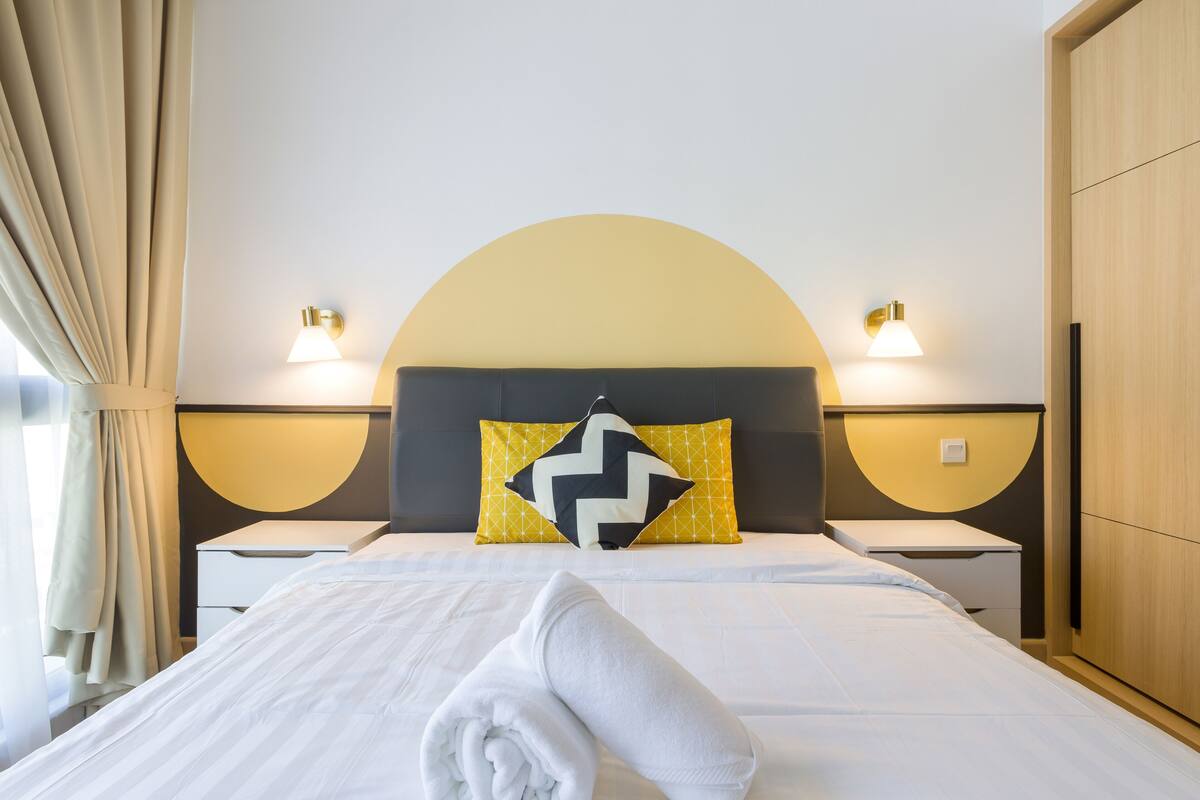 Een net opgemaakt bed met een opgerolde handdoek, een geel-zwart kussen en twee nachtkastjes met lampen.