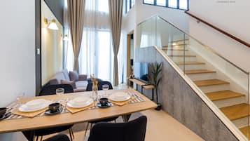 A modern dining area with a wooden table set for two, a sofa, and a staircase with glass railings.