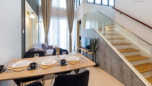 A modern dining area with a wooden table set for two, a sofa, and a staircase with glass railings.