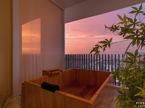 A wooden bathtub in a room with a large window overlooking a cityscape at sunset.