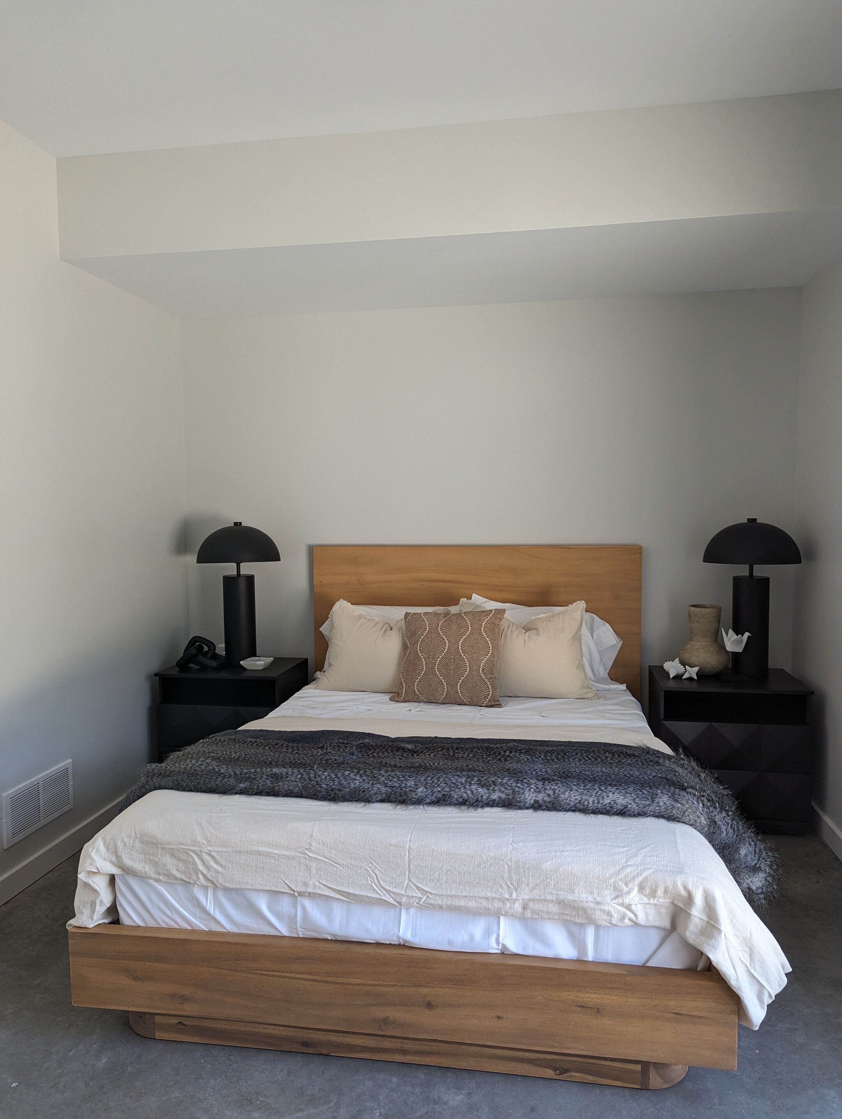 Un dormitorio con cama de madera, ropa de cama blanca, dos mesitas de noche con lámparas y una manta gris.