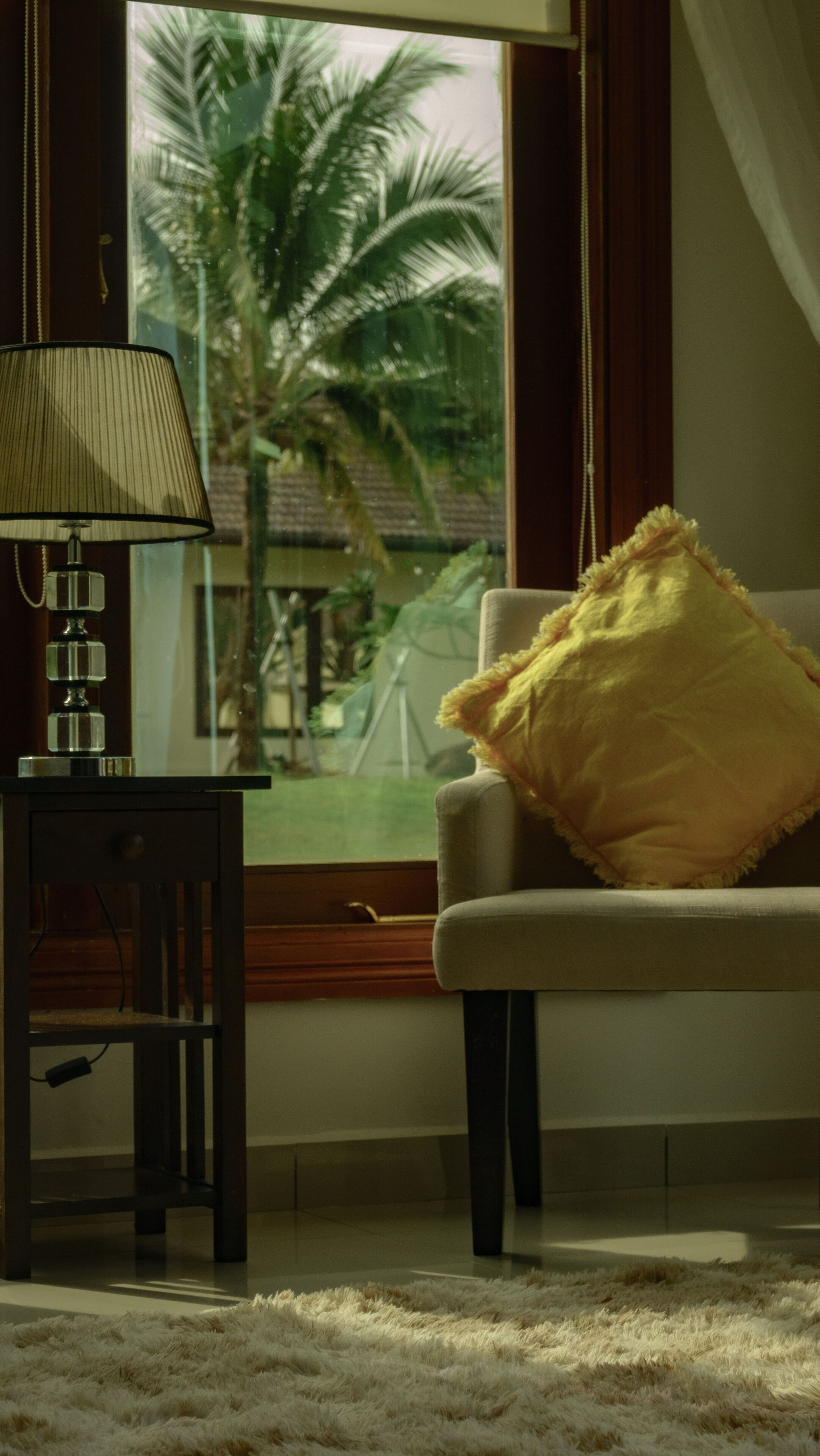 A room with a lamp on a side table, a chair with a yellow pillow, and a view of palm trees through a window.