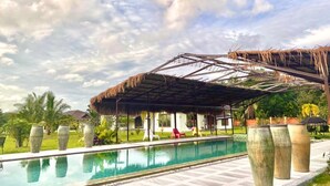 Ein Pool mit einer strohgedeckten Struktur, umgeben von großen Pflanzgefäßen und Palmen.