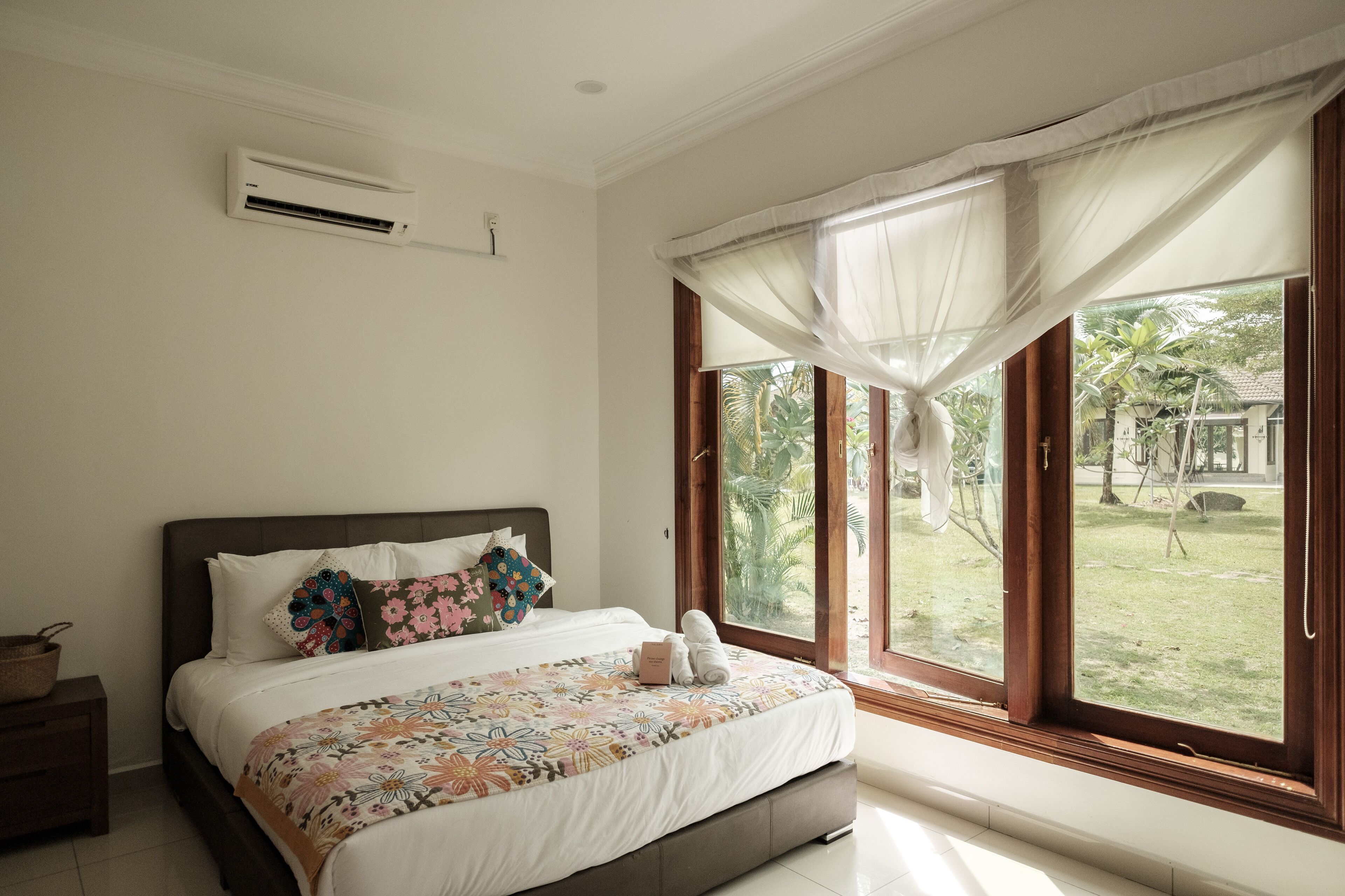 A bedroom with a large bed, a window with sheer curtains, and an air conditioning unit on the wall.