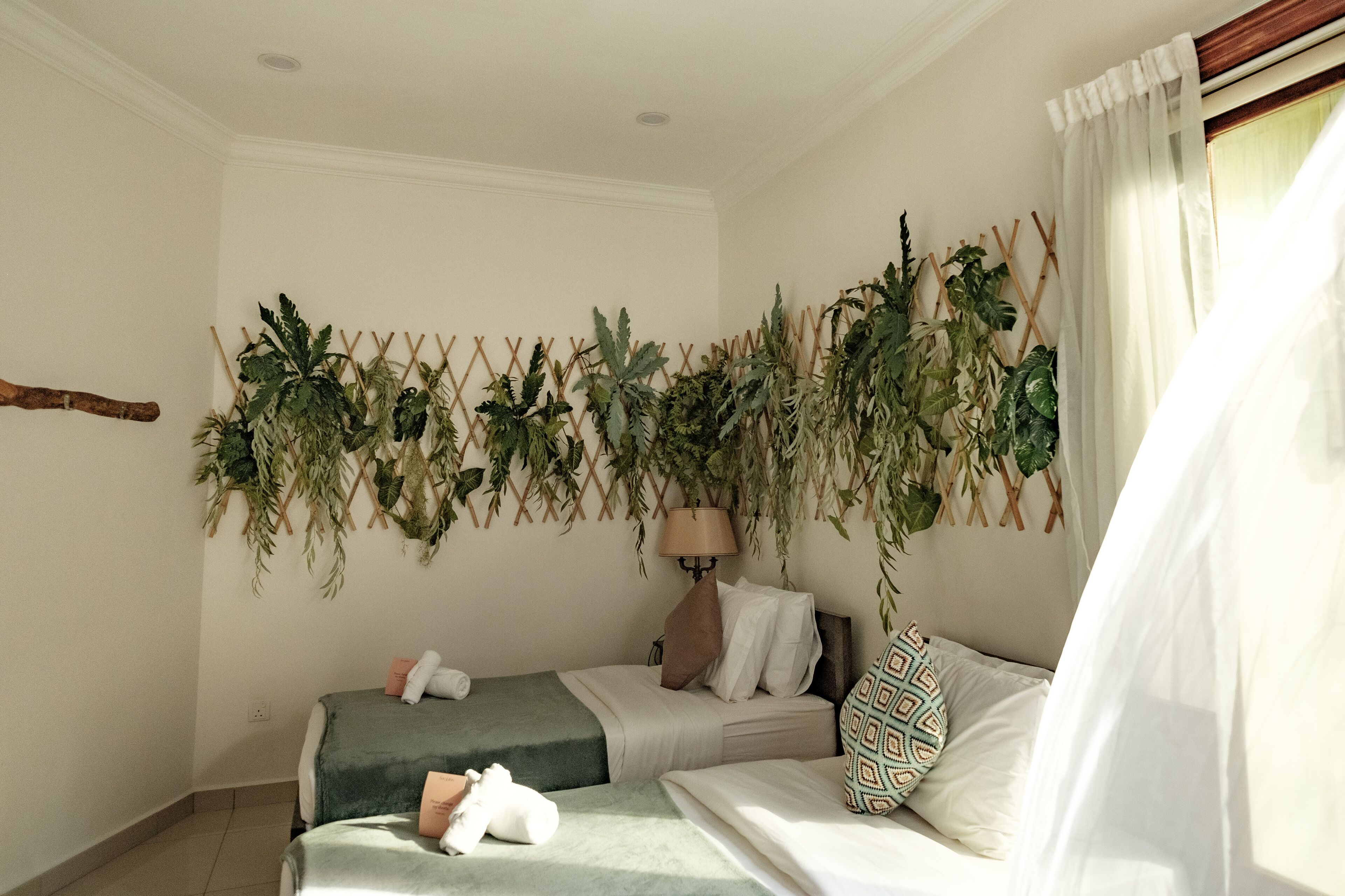Un dormitorio con dos camas, un arreglo de plantas en la pared, una lámpara de noche y una ventana con cortinas.