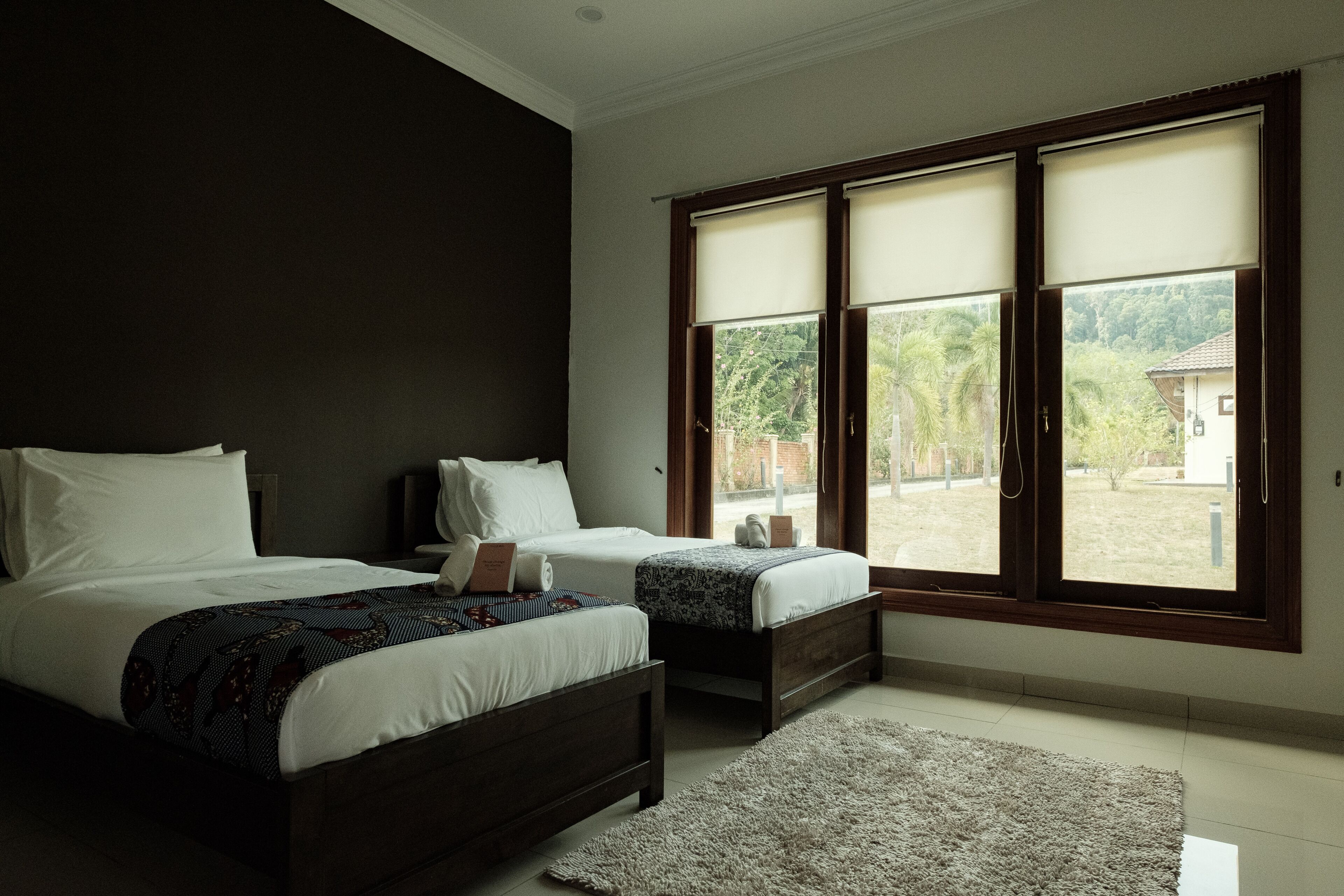 A bedroom with two beds, a large window with blinds, and a view of greenery outside.