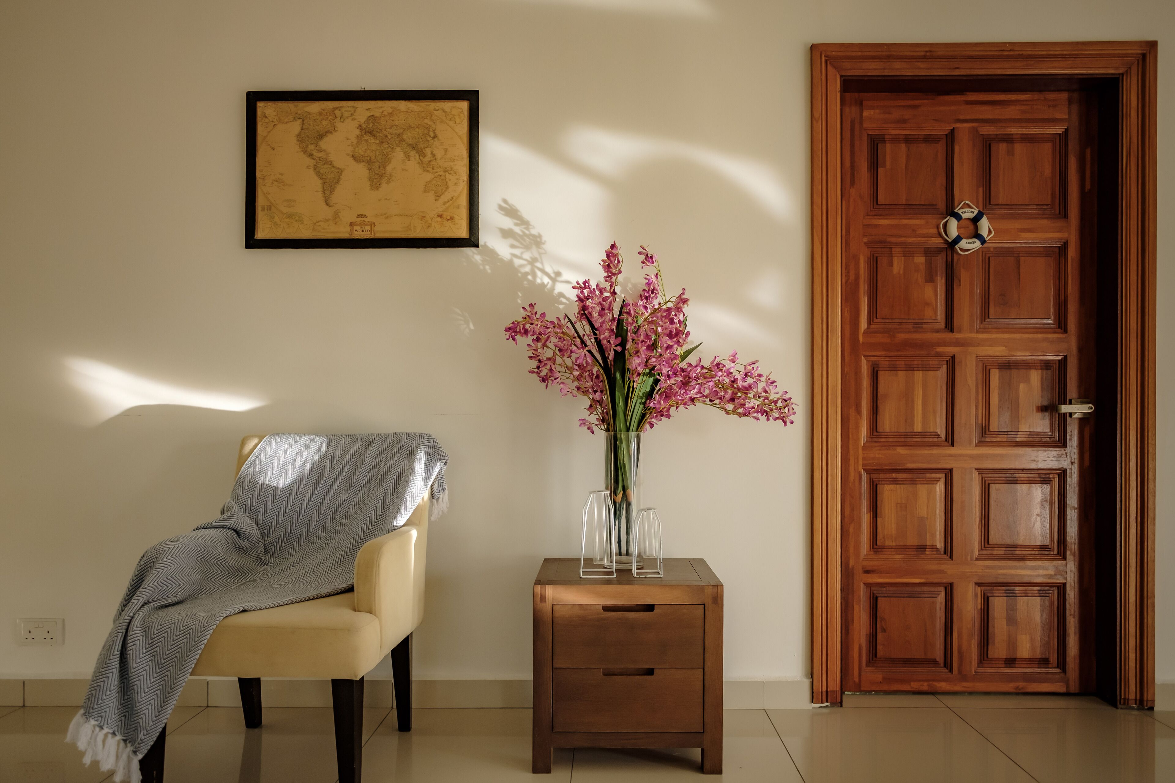A room with a wooden door, a map on the wall, a chair with a blanket, and a vase with flowers on a side table.
