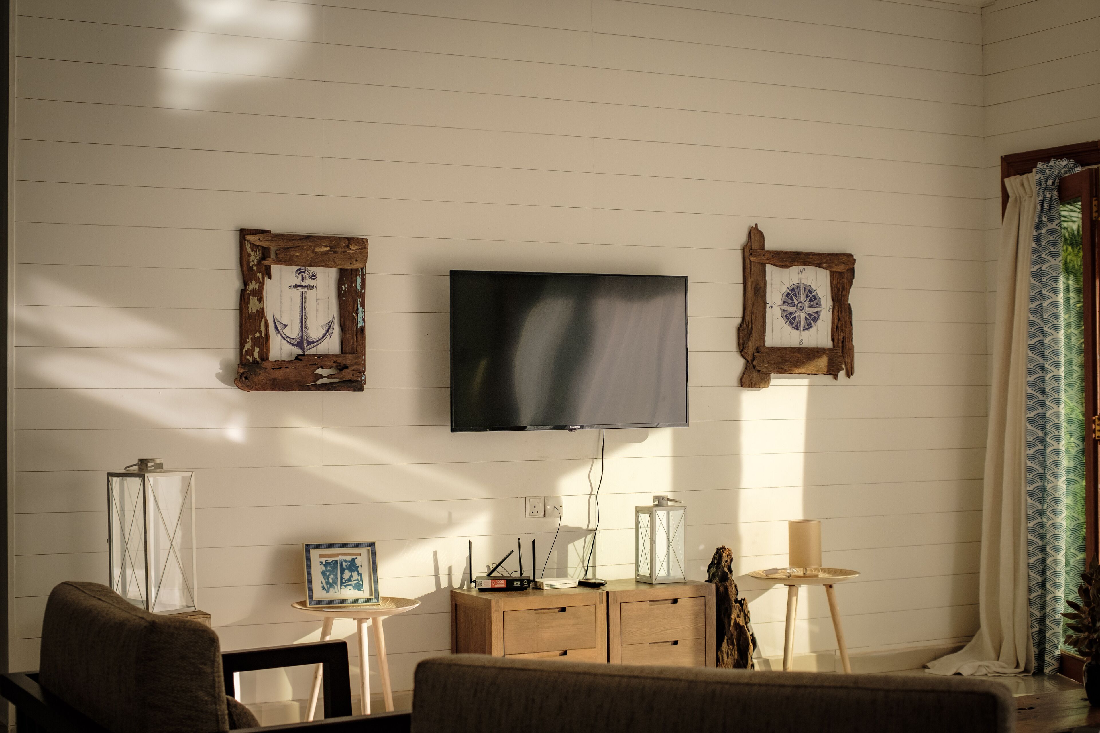 A living room with a flat-screen TV mounted on the wall, wooden furniture, and a window with patterned curtains.