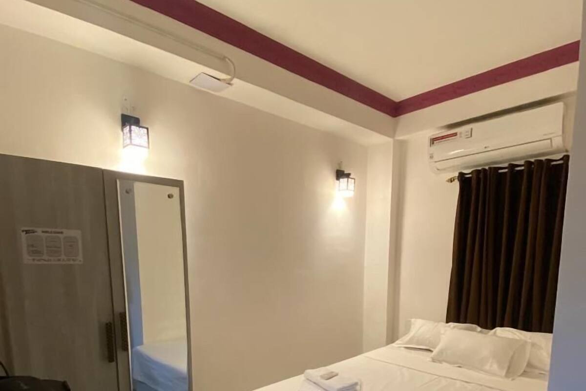 A hotel room with a bed, a desk with a chair, a mirror, and an air conditioning unit.