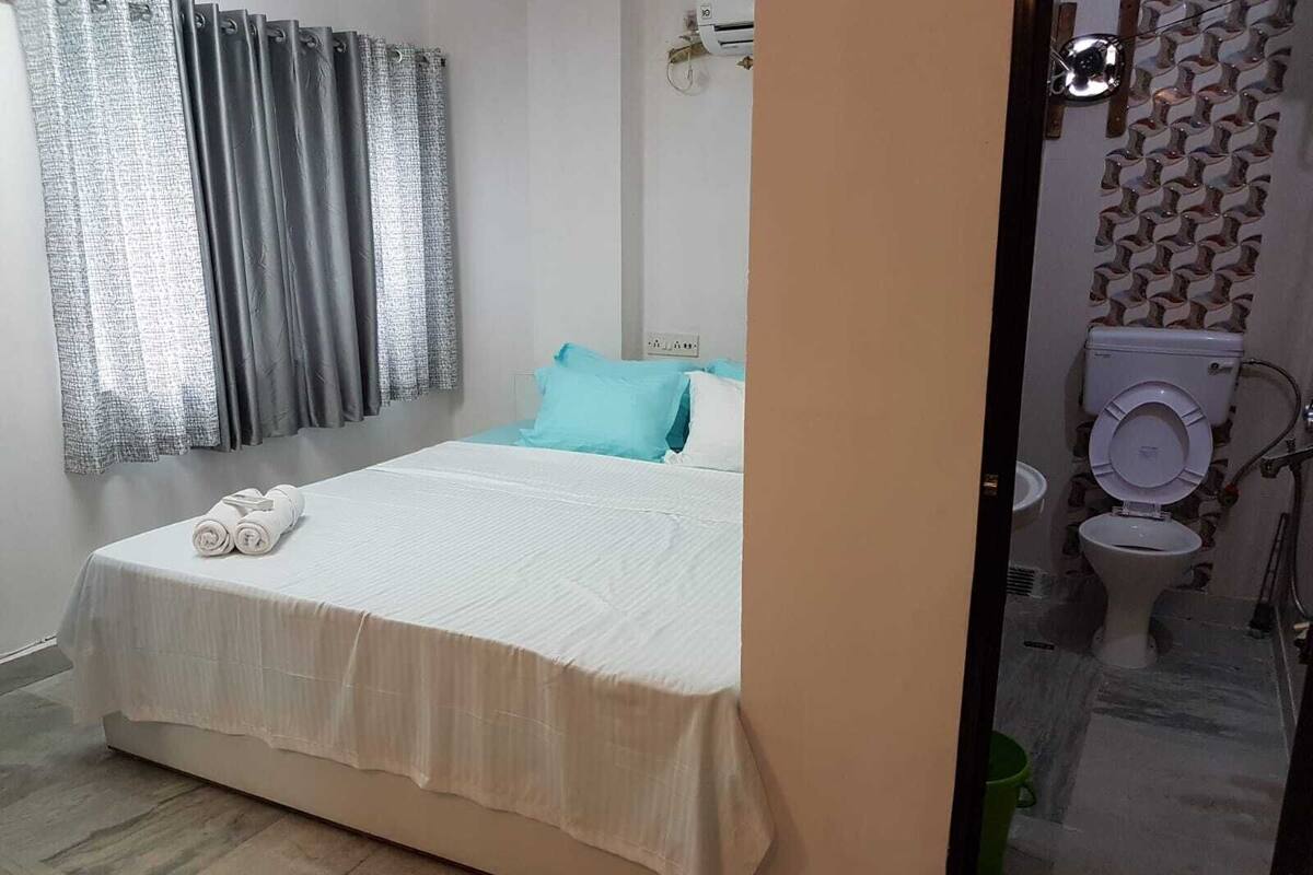 A bedroom with a bed, pillows, a window with curtains, and a bathroom visible through a glass door.