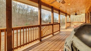 A covered porch with wooden beams and a ceiling fan, overlooking a forested area.