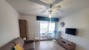A living room with a sofa, a TV, a ceiling fan, and a sliding glass door leading to an outdoor area. - Renovated studio close to the beach of Sainte Asile in Pin Rolland (ST MANDRIER SUR MER)