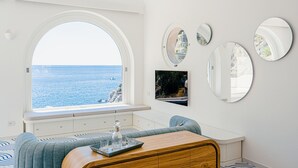 Een kamer met een groot boogvenster met uitzicht op zee, een zithoek met een bank en een tafel, en verschillende spiegels aan de muur.
