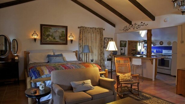 A cozy room with a bed, a sofa, a small table, and a chair. There is a kitchen area visible in the background. - Private Cottage “Storybook Barn” (Julian)