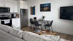 Cozinha moderna com armários brancos, geladeira de aço inoxidável e micro-ondas. Há uma área de jantar com mesa e cadeiras, e um quadro com uma paisagem na parede.