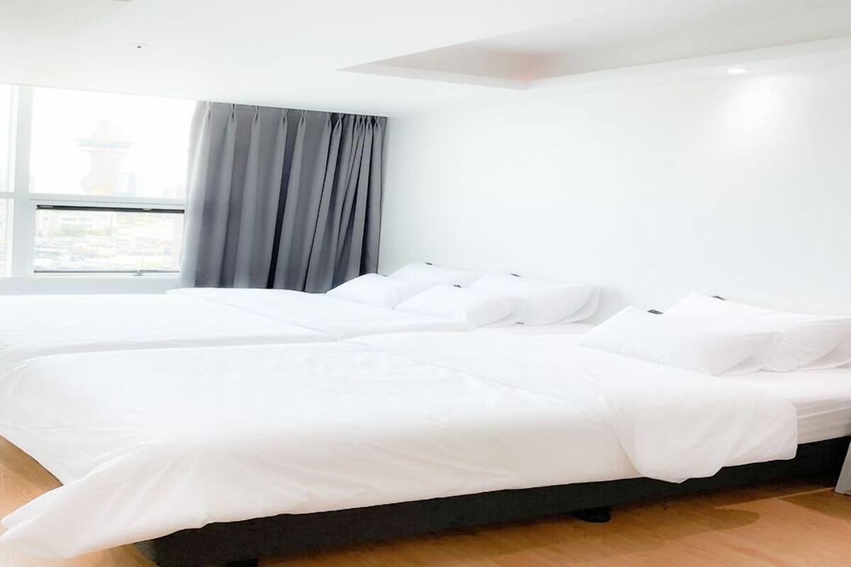 A modern hotel room with two single beds, a large window with a view of a cityscape, and a grey curtain.