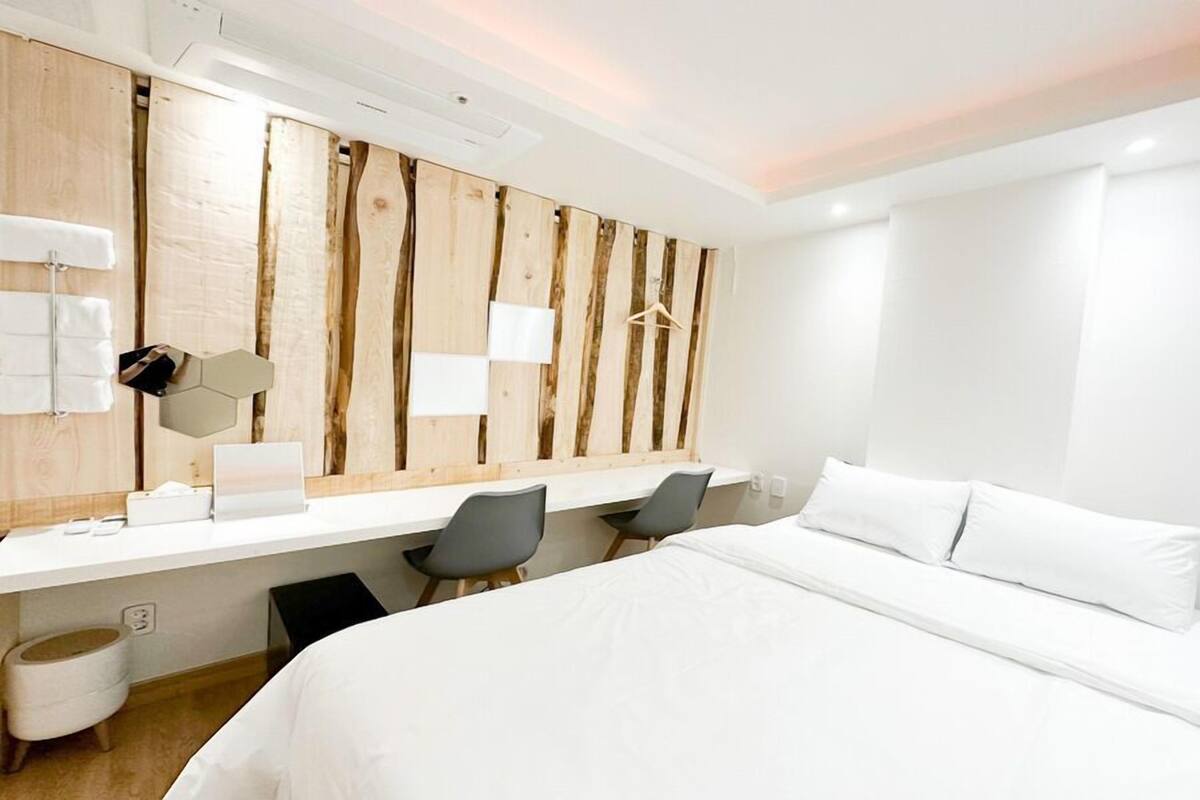 A modern hotel room with a large bed, a desk with a chair, a wooden accent wall, and a large mirror.