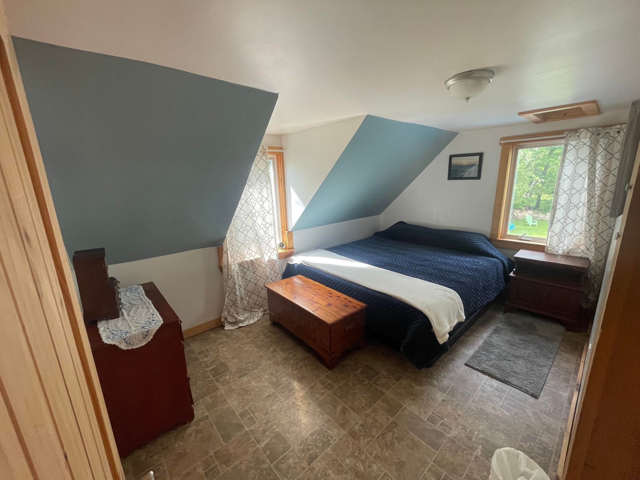 Une chambre avec un plafond en pente, un lit recouvert d’une couvre-lit à motifs, un banc en bois, une petite table et une fenêtre avec des rideaux.