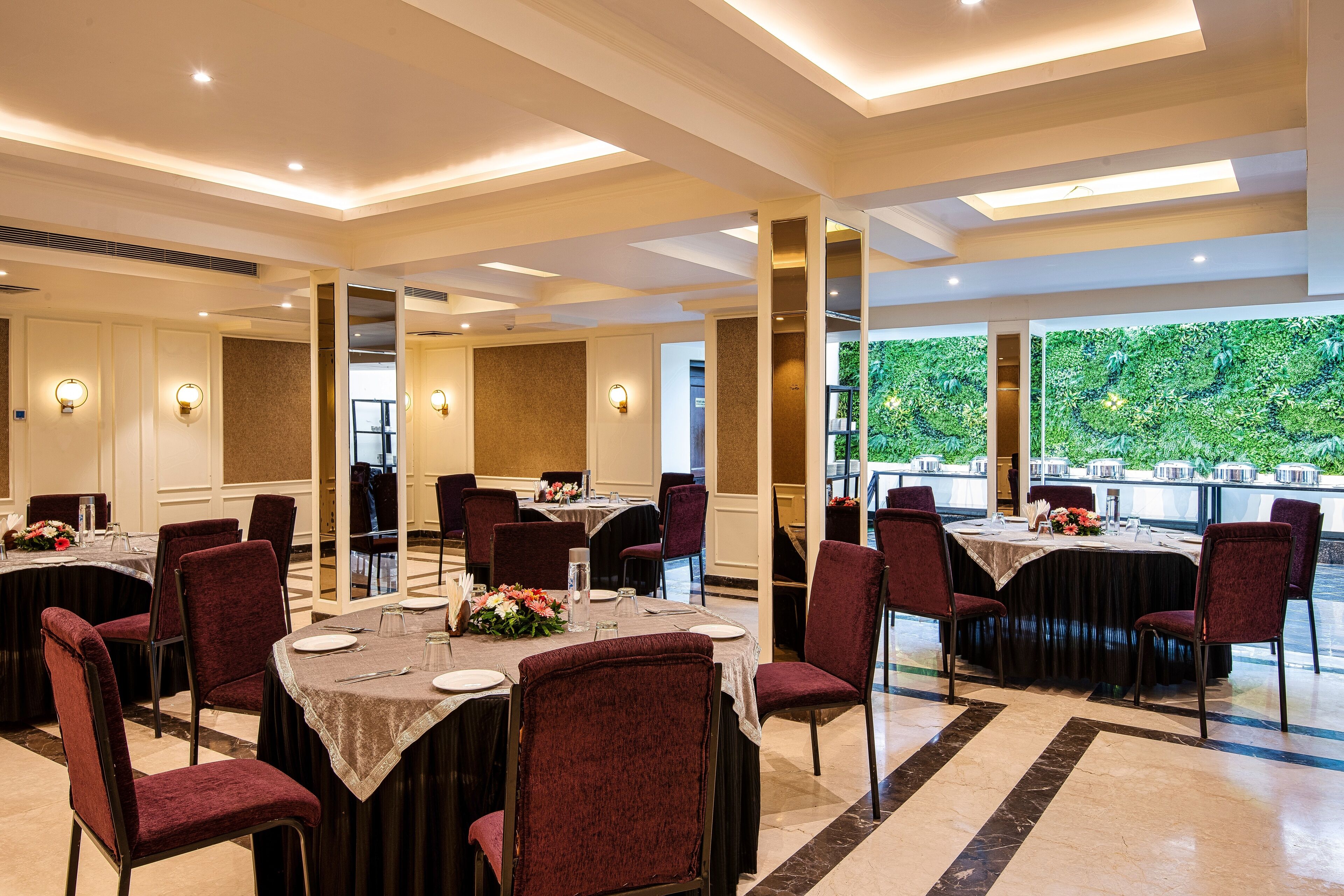 A hotel dining area with round tables, red upholstered chairs, and a view of greenery through large windows.
