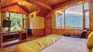 Een houten kamer met een bed, een bureau, een televisie en een groot raam met uitzicht op de bergen.