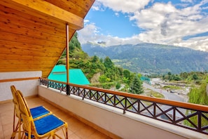 A balcony with a wooden ceiling, two wicker chairs with blue cushions, and a view of a mountainous landscape with a green-roofed building in the distance. - Solang Cottage By Snow City Farm (Manali)