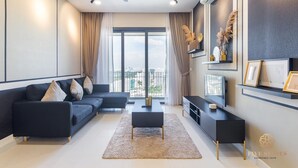 Ein modernes Wohnzimmer mit einem dunklen Sofa, einem Couchtisch und einem Fernseher auf einem schwarzen Lowboard. Es gibt ein großes Fenster mit Vorhängen, einen Deckenventilator und ein wandmontiertes Regal mit Dekorationen.