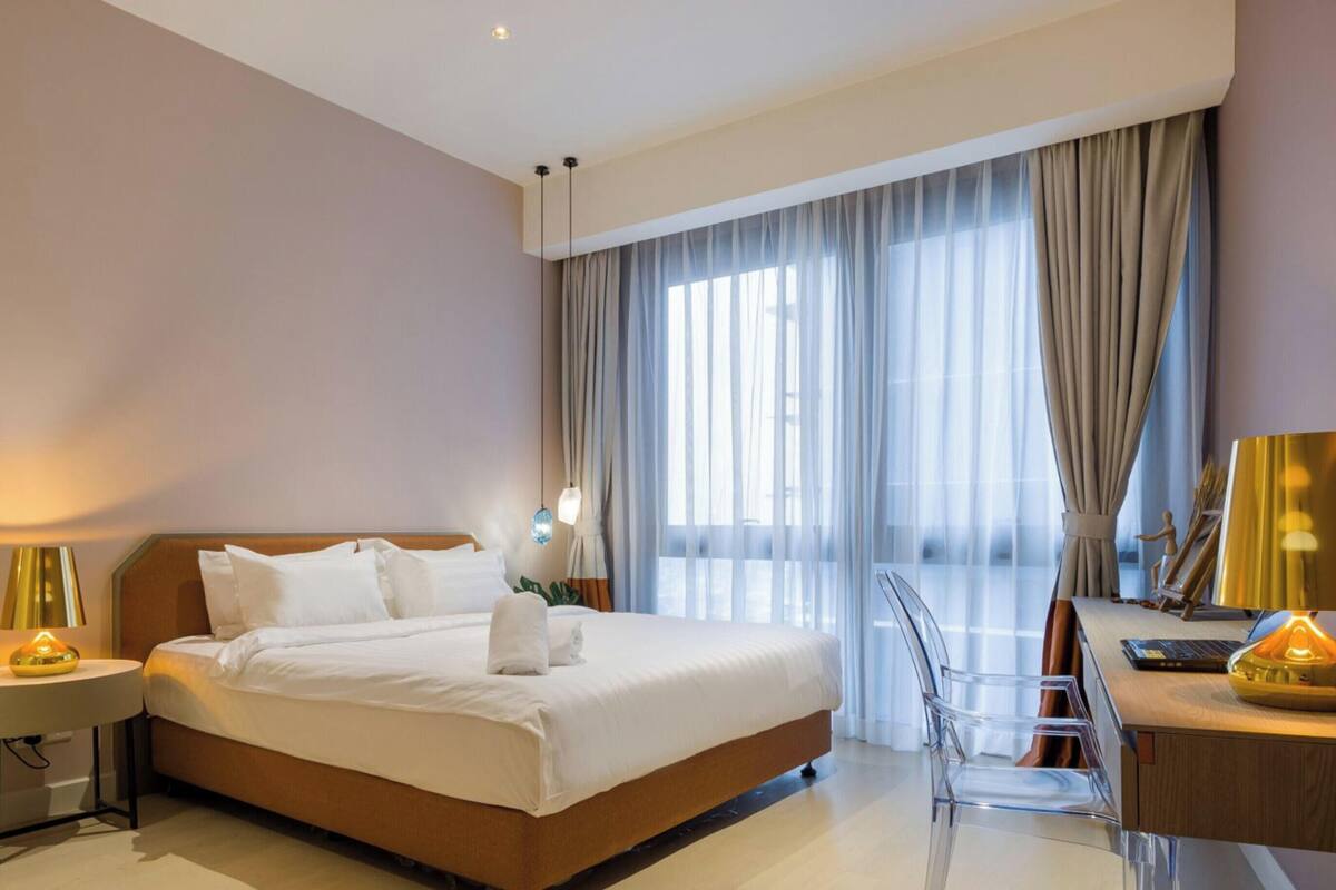 Quarto de hotel com cama, escrivaninha, cadeira, luminária e janela com cortinas.