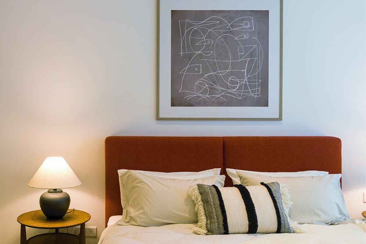 Uma cama bem arrumada com roupa de cama branca e almofadas listradas, um criado-mudo redondo de madeira com uma luminária e uma obra de arte abstrata emoldurada na parede.