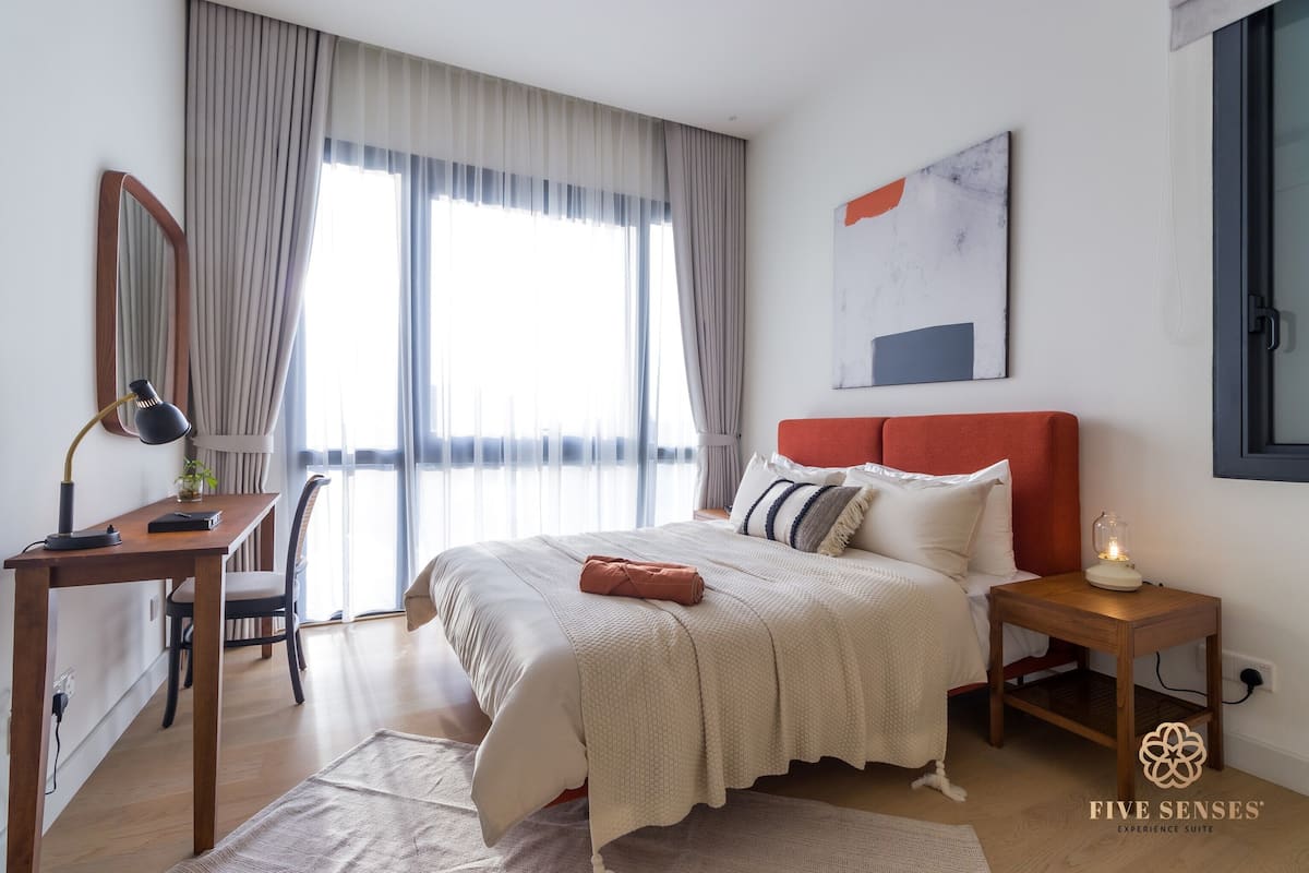 Quarto com cama, mesa de cabeceira, luminária e uma grande janela com cortinas.