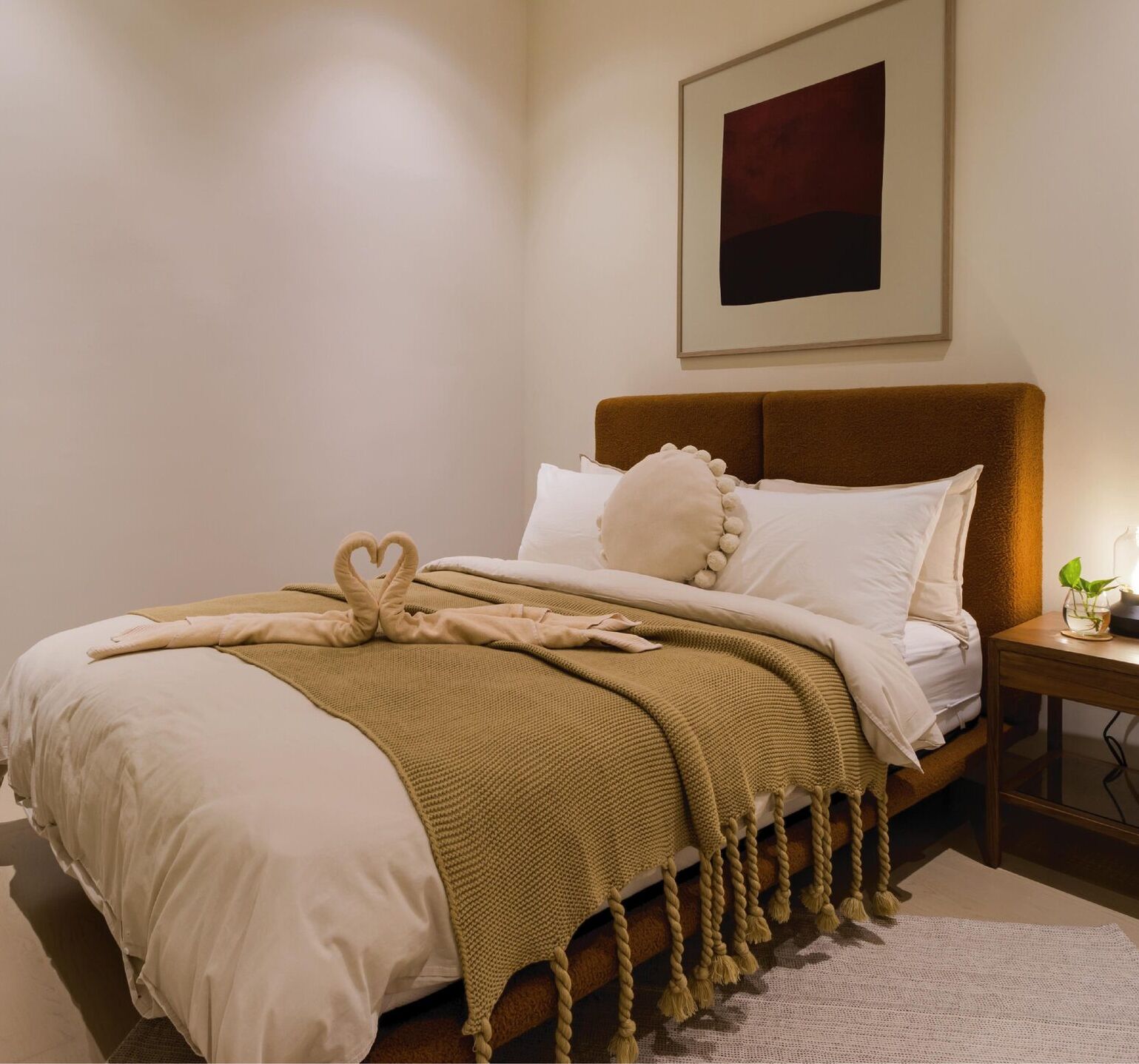 A neatly made bed with a brown headboard, a beige throw blanket, and a heart-shaped pillow.