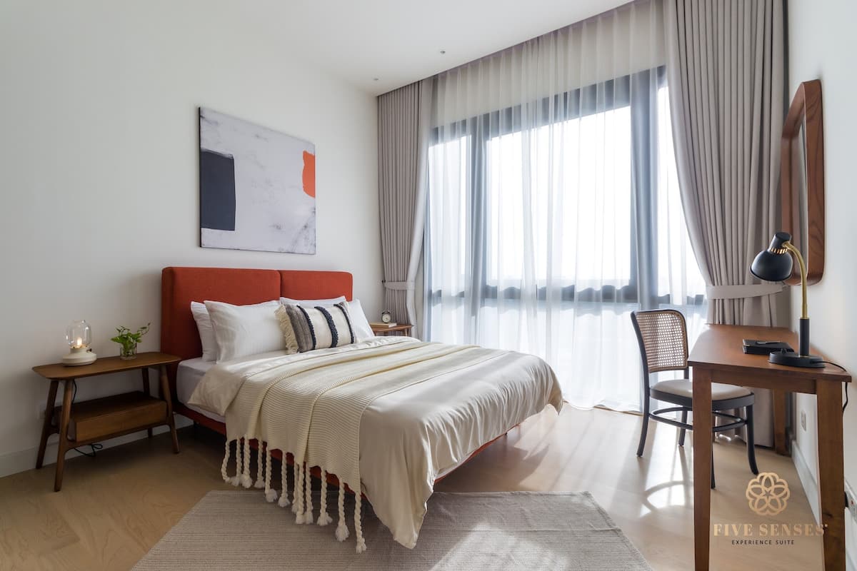 Quarto moderno com uma cama grande, uma escrivaninha e uma cadeira. Há uma janela com cortinas, uma mesinha de cabeceira com luminária, e uma obra de arte na parede.