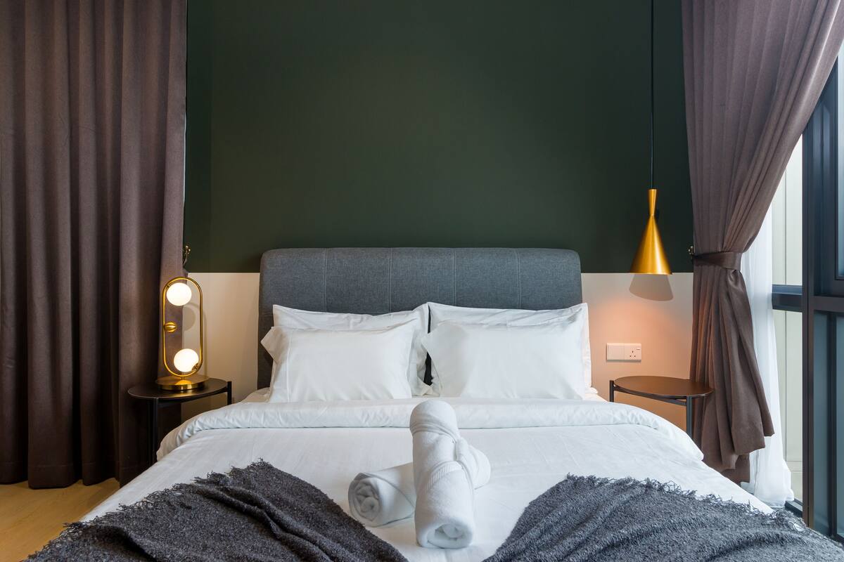 Cama bem arrumada com roupa de cama branca, uma toalha dobrada e um cobertor cinza escuro. Há duas mesas de cabeceira, uma com abajur e outra com tomada. O quarto tem paredes verde escuro e cortinas marrons.