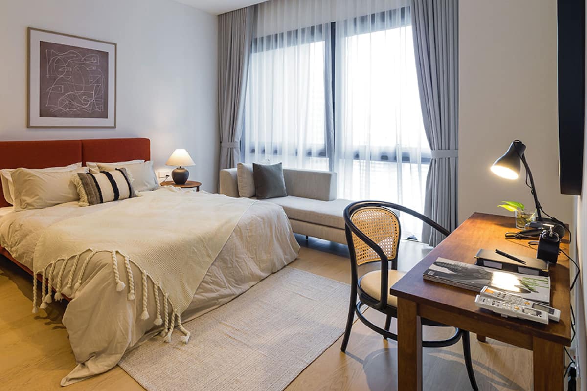 Quarto de hotel com cama, mesas de cabeceira, escrivaninha com cadeira, sofá e uma janela grande com cortinas.