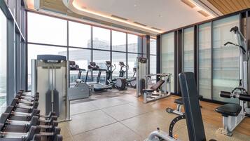 A modern gym with various exercise equipment, large windows, and a high ceiling.