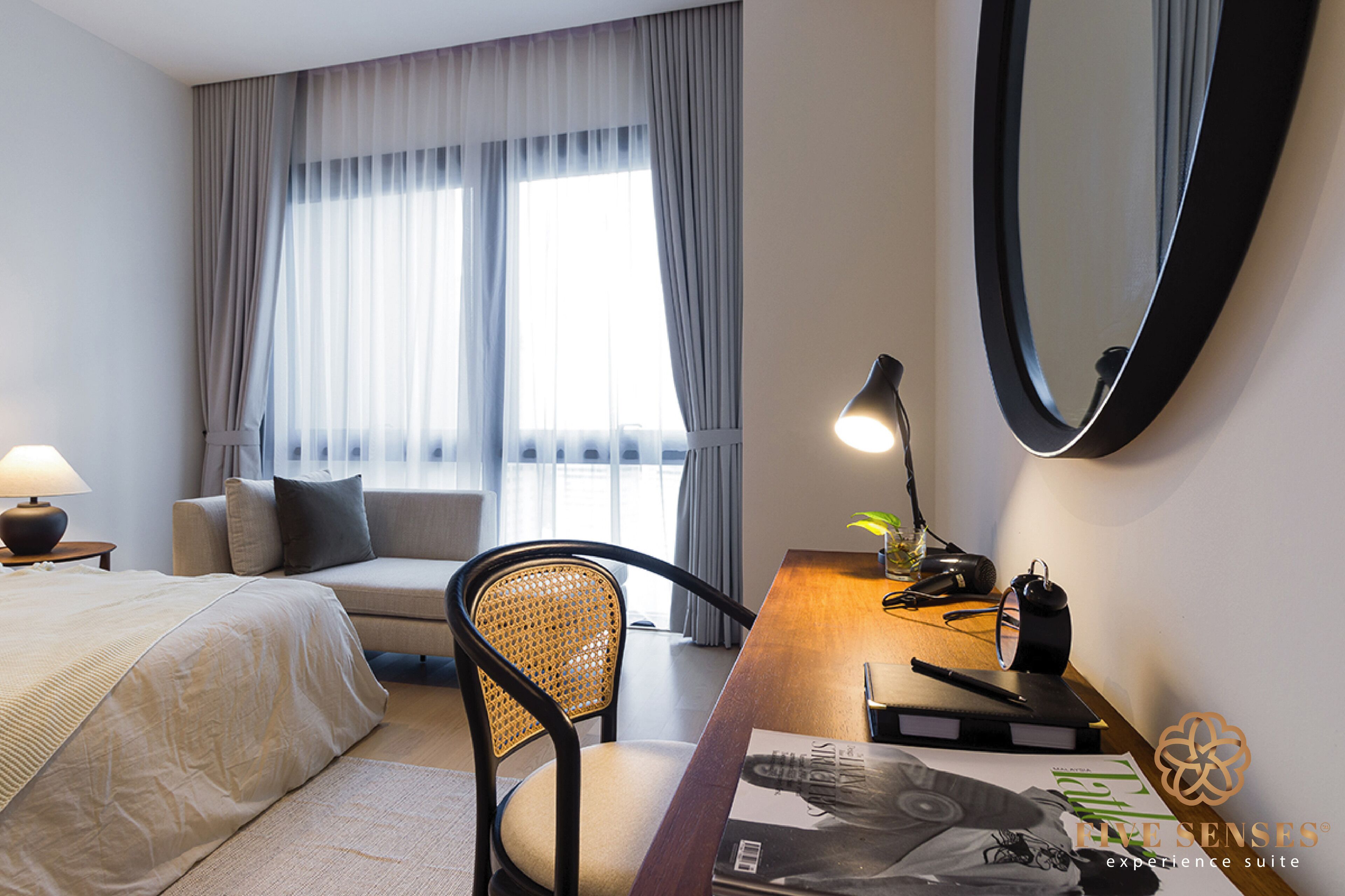 A hotel room with a bed, a desk with a lamp, a chair, a large mirror, and a window with curtains.