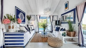 A spacious living room with a white ceiling, large windows, and a view of the sea. There are two white sofas, a coffee table, and a round ottoman. - Myrtle Cottage - Charming 2 bed Cottage (Paxos)