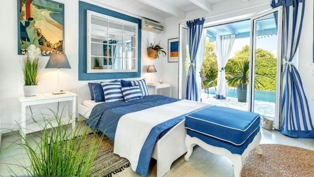 A bedroom with a large bed, a window with curtains, a bedside table with a lamp, and a view of greenery outside. - Myrtle Cottage - Charming 2 bed Cottage (Paxos)